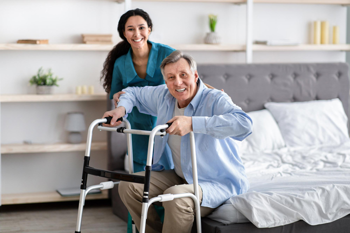 Female nurse helping elderly male with walking frame stand up from bed at home