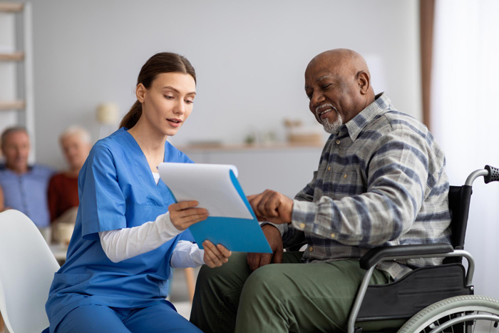 Young woman in blue workwear nurse helping senior black man in wheelchair with questionnaire